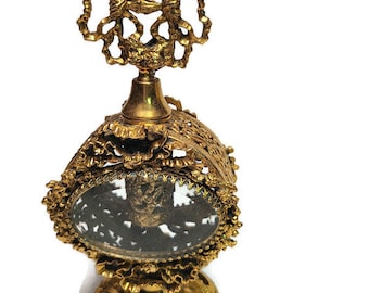 May include: A gold-toned perfume bottle with a decorative, ornate design. The bottle has a clear glass center and a detailed, intricate top.