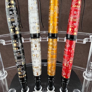 May include: Four ballpoint pens with circuit board designs. The pens are black, white, gold, and red, each with a black clip and tip. They are displayed in a clear acrylic stand with labeled slots.