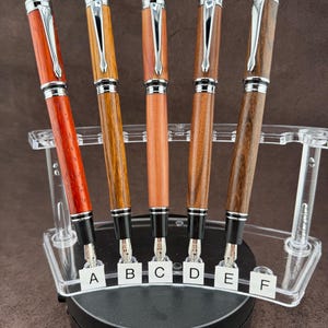 May include: A collection of six fountain pens with wooden barrels in various shades of brown and red. Each pen has a silver-colored clip and accents. The pens are displayed in a clear acrylic stand, with labels A through F.