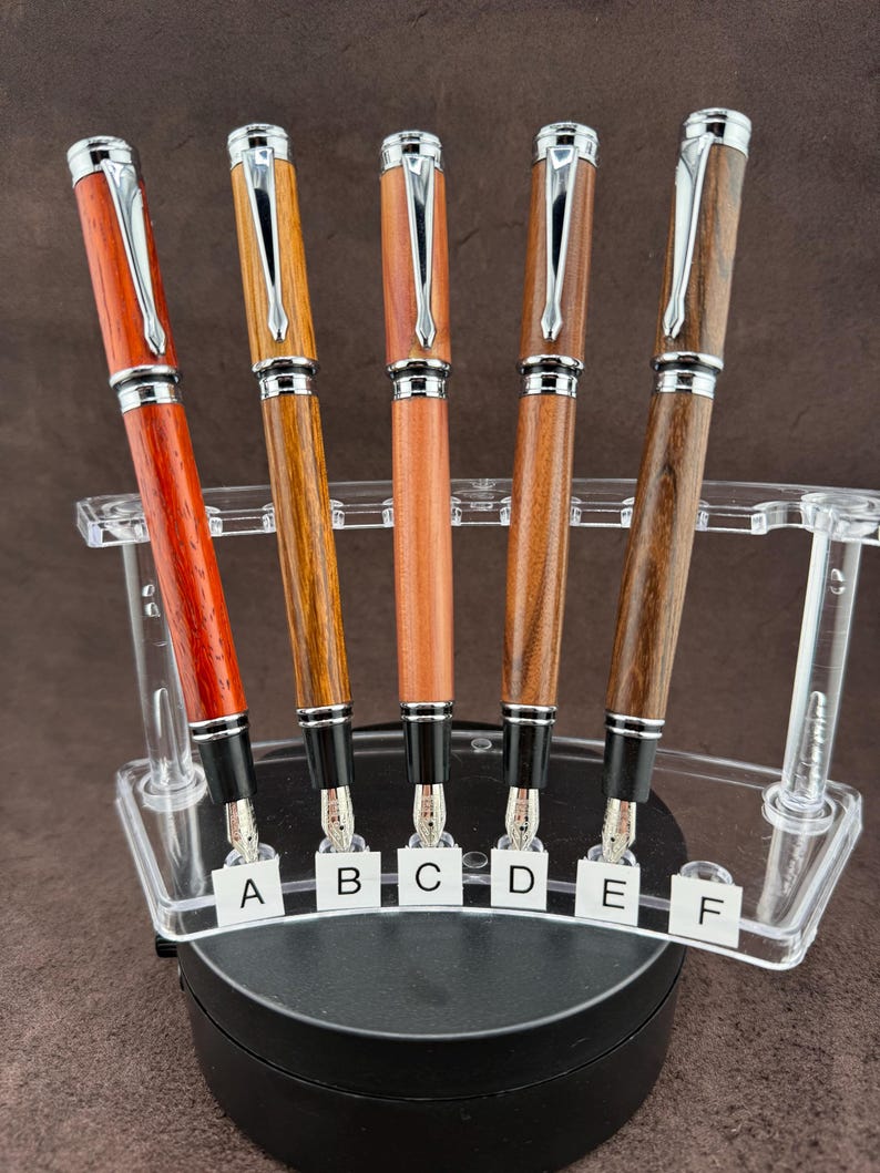 May include: A collection of six fountain pens with wooden barrels in various shades of brown and red. Each pen has a silver-colored clip and accents. The pens are displayed in a clear acrylic stand, with labels A through F.