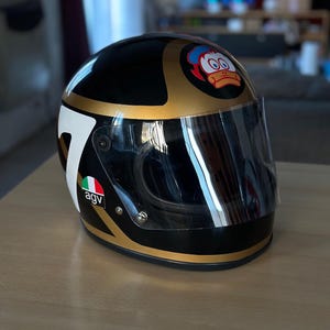 Könnte beinhalten: Ein schwarz-goldener Motorradhelm mit klarem Visier, einer großen weißen Nummer sieben an der Seite, einem AGV-Logo in den Farben der italienischen Flagge und einer kreisförmigen Grafik einer Cartoon-Ente im vorderen oberen Bereich der Schale.