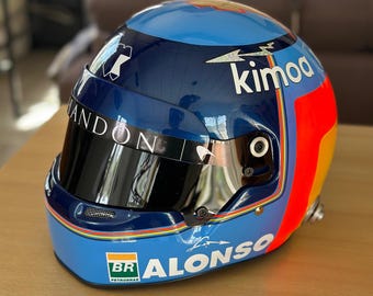 May include: A blue and navy racing helmet with the name "Alonso" and the word "kimoa" printed on it. The helmet has a black visor and a multi-colored stripe design. The helmet is sitting on a wooden surface.