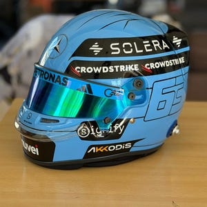 Può includere: Un casco da corsa azzurro chiaro con visiera a specchio. Il casco presenta i loghi di "SOLERA", "CROWDSTRIKE", "PETRONAS", "Signify" e "AKKODIS". Il numero "63" è visibile. Il casco è su una superficie di legno.