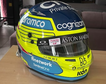 May include: A racing helmet with a yellow and blue design. The helmet features the logos of "Aramco", "Cognizant", "Aston Martin", and "finetwork SentinelOne". The helmet also has the text "B&O crypto.com icon".