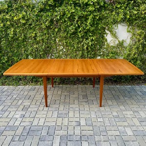 May include: A rectangular wooden dining table with a warm, light brown finish and visible wood grain. The table features a flat, smooth surface with a central seam indicating an extendable design, supported by four tapered, angled wooden legs.