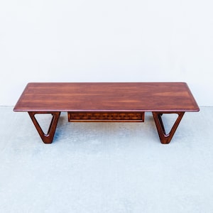 May include: A mid-century modern coffee table with a rectangular top and distinctive V-shaped legs. The table is made of dark wood and features a small drawer with a woven design. The table measures approximately 48 inches long.