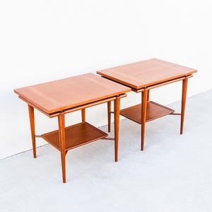 May include: Two matching mid-century modern side tables. Each table has a rectangular top with a lighter wood border, a lower shelf, and tapered legs. The tables are made of wood and have a warm, reddish-brown finish.