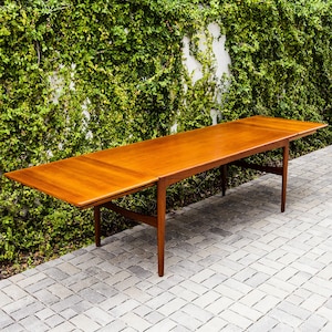 May include: A long, rectangular wooden table with a warm, reddish-brown finish. The table has a leaf extension on each end, and is supported by slender, angled legs. The table is set on a gray brick patio, with a green wall of foliage in the background.