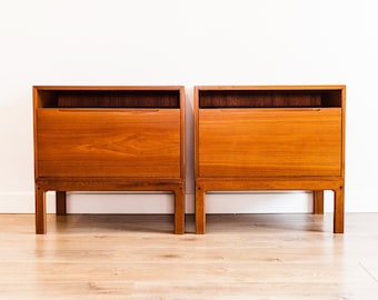 May include: Two matching wooden nightstands with a warm, reddish-brown finish. Each nightstand has a drawer and an open shelf at the top. The nightstands are raised on four legs and are set against a white wall.
