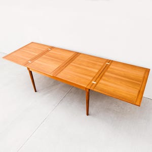 Puede incluir: Una mesa de comedor de madera marrón claro con una tapa rectangular y cuatro patas delgadas y cónicas. La mesa tiene una extensión de hoja en cada extremo, y la veta de la madera es visible. La mesa mide aproximadamente 183 cm de largo cuando está completamente extendida.