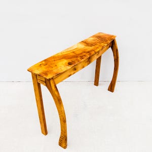 Puede incluir: Una consola de madera hecha a mano con un acabado natural. La mesa presenta una tapa rectangular y patas curvas, mostrando la veta y los nudos de la madera. Los tonos cálidos marrón dorado de la madera crean una estética rústica y elegante. Un mueble único.