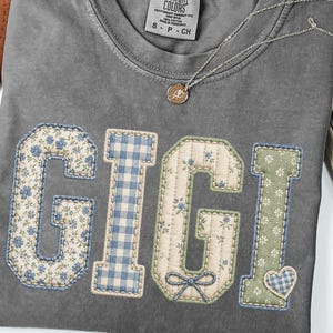 Puede incluir: Una camiseta de manga corta gris carbón con la palabra GIGI en letras grandes aplicadas de tela. Las letras tienen patrones variados que incluyen flores azules, cuadros vichy azules, tela acolchada color crema con un lazo y flores verdes.