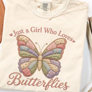 Puede incluir: Una camiseta de manga corta color crema con un gráfico central de una mariposa estilo patchwork en tonos apagados de rosa, verde y azul. El texto "Just a Girl Who Loves Butterflies" está impreso en una fuente cursiva sobre y bajo la mariposa.