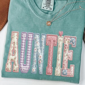 May include: A sage green short-sleeve t-shirt with a crew neckline and a Comfort Colors brand label. The front features the word AUNTIE in large varsity-style letters, each filled with various floral and gingham fabric patterns, plus a small heart.