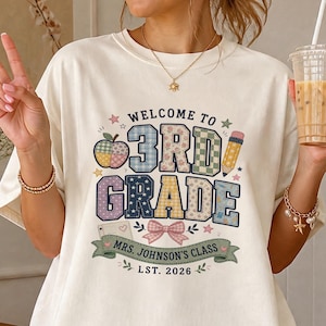 Peut inclure: Un t-shirt crème arborant un motif avec le texte "WELCOME TO 3RD GRADE" dans un style patchwork, un crayon, une pomme et un nœud. Une bannière verte en dessous indique "MRS. JOHNSON'S CLASS" et "LST. 2026" avec de petites étoiles.