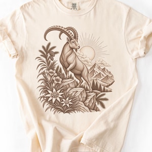 Puede incluir: Una camiseta color marfil con un gráfico en tinta marrón que muestra un íbice sobre unas rocas. La ilustración incluye picos de montañas, un sol radiante, pinos y flores de edelweiss. La camiseta tiene cuello redondo y mangas cortas.