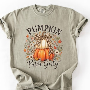 Puede incluir: Una camiseta de manga corta color verde salvia con un gráfico de dos calabazas apiladas coronadas por un lazo con estampado de leopardo. El diseño está rodeado de hojas y flores otoñales, con el texto PUMPKIN Patch Girly impreso en negro.