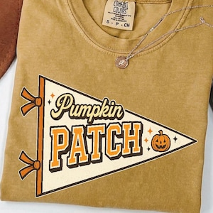 Puede incluir: Una camiseta de manga corta color amarillo mostaza con un gráfico de banderín triangular color crema que presenta el texto "Pumpkin PATCH" en letras marrones y naranjas, junto a un pequeño icono de calabaza naranja y dos cintas naranjas.