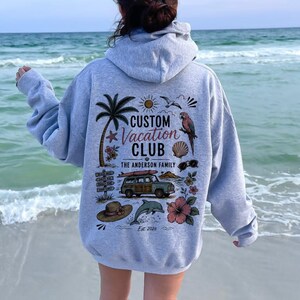 May include: A light gray hooded sweatshirt featuring a back graphic with the text CUSTOM VACATION CLUB THE ANDERSON FAMILY EST. 2026. The design includes a palm tree, a parrot, a station wagon with a surfboard, a dolphin, a sun, and flowers.