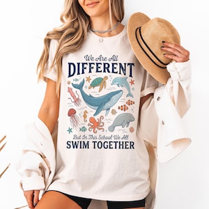 Puede incluir: Camiseta color crema con un gráfico de vida marina que incluye una ballena, un pulpo, una medusa y otras criaturas marinas. El texto de la camiseta dice: "We Are All Different But In This School We All Swim Together."