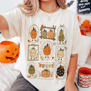 Puede incluir: Una camiseta de color crema con un estampado gráfico de varias calabazas ilustradas en diferentes patrones y colores, incluyendo naranja, verde y negro, dispuestas en recuadros con pequeños detalles florales y bordes decorativos.
