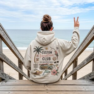 May include: A beige hooded sweatshirt featuring a back graphic with a palm tree, a parrot, a station wagon with a surfboard, a dolphin, a sun, a seashell, a hat, and flowers. The text reads Custom Vacation Club, The Anderson Family, and Est. 2026.