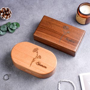 May include: Two wooden boxes, one rectangular and one oval, with floral engravings and personalized names. The rectangular box is a dark brown, while the oval box is a lighter wood tone. Both boxes have a smooth, polished finish.