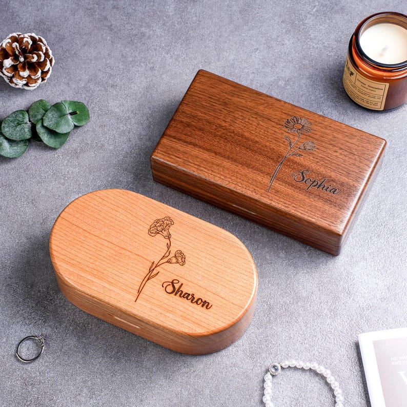 May include: Two wooden boxes, one rectangular and one oval, with floral engravings and personalized names. The rectangular box is a dark brown, while the oval box is a lighter wood tone. Both boxes have a smooth, polished finish.