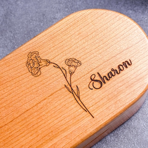 May include: A light-colored wooden box with rounded edges. The top is engraved with a floral design and the name "Sharon" in cursive script. The wood grain is visible, and the box appears to be made of a light-colored hardwood.