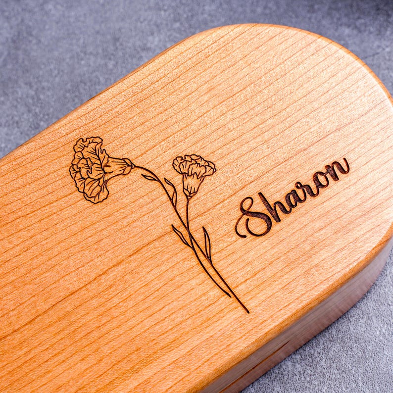 May include: A light-colored wooden box with rounded edges. The top is engraved with a floral design and the name "Sharon" in cursive script. The wood grain is visible, and the box appears to be made of a light-colored hardwood.