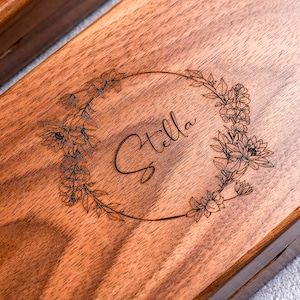 May include: Close-up of a wooden box with a floral wreath design engraved on the lid. The word "Stella" is elegantly scripted within the wreath. The wood has a warm, natural tone, and the engraving is dark.