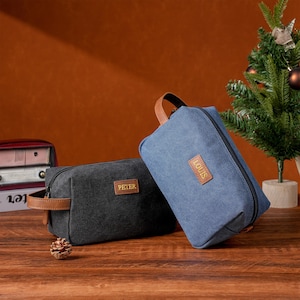 May include: Two canvas toiletry bags, one black and one blue, with brown leather accents. The black bag has "PETER" on a leather patch, and the blue bag has "LOUIS". Both have a zipper closure and a handle.
