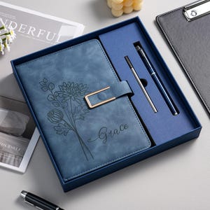May include: A blue notebook with a floral line art design and the name Grace printed on the cover, featuring a gold-toned rectangular magnetic closure. The notebook is presented in a matching blue gift box alongside a blue pen and a silver ink refill.