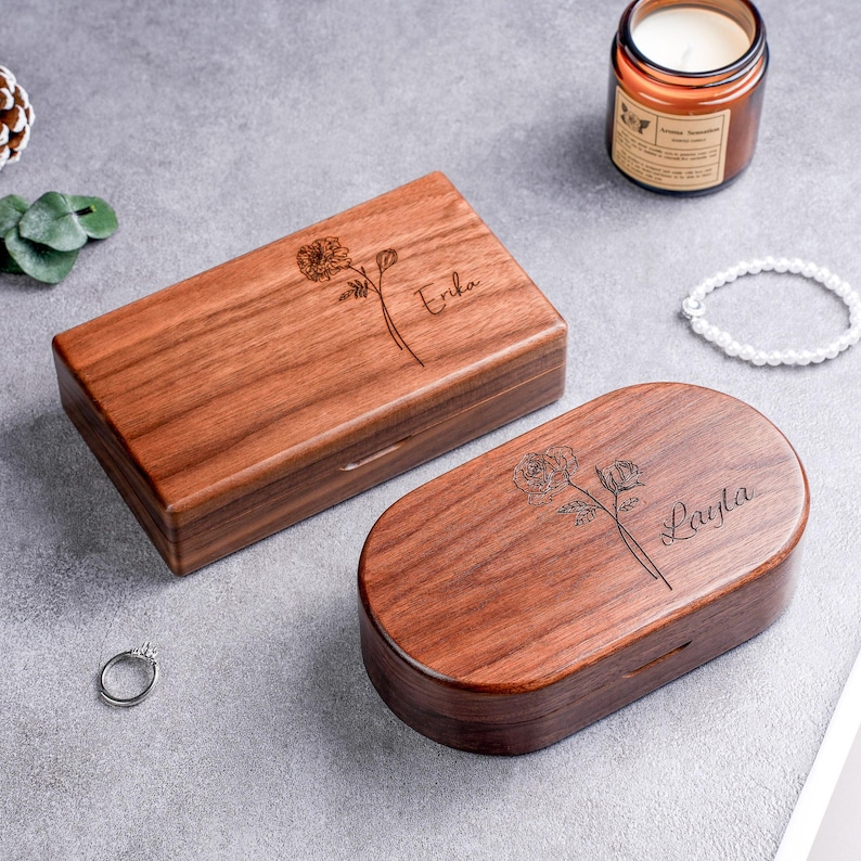 May include: Two wooden jewelry boxes, one rectangular and one oval, with floral engravings and personalized names. The rectangular box is labeled "Erika," and the oval box is labeled "Layla." Both boxes are made of a warm brown wood.