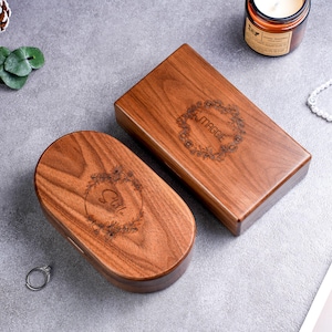 May include: Two wooden jewelry boxes with engraved floral designs and names. One box is rectangular and the other is oval. The rectangular box has the name "Maggie" and the oval box has the name "Stella". The boxes are made of a warm brown wood.