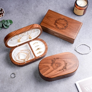 May include: Wooden jewelry boxes with white interiors, holding necklaces, bracelets, and rings. The boxes are oval and rectangular, with floral engravings and names on the lids. A candle and a bracelet are also visible.