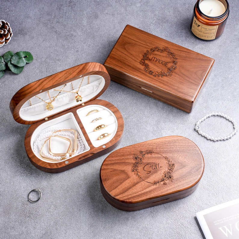 May include: Wooden jewelry boxes with white interiors, holding necklaces, bracelets, and rings. The boxes are oval and rectangular, with floral engravings and names on the lids. A candle and a bracelet are also visible.