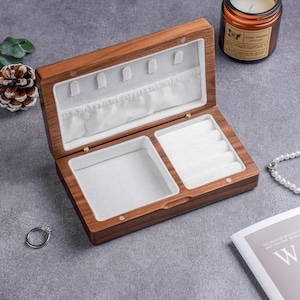 May include: Open wooden jewelry box with a light-colored interior. The box is rectangular with multiple compartments, including ring holders and a larger space. The wood has a warm, natural tone. A ring and a pearl necklace are nearby.