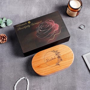 May include: A rectangular black gift box featuring a red rose graphic and the text "Moments, Beautifully Wrapped." alongside an oval wooden box lid engraved with the name "Rachel" inside a circular floral wreath design on a gray surface.