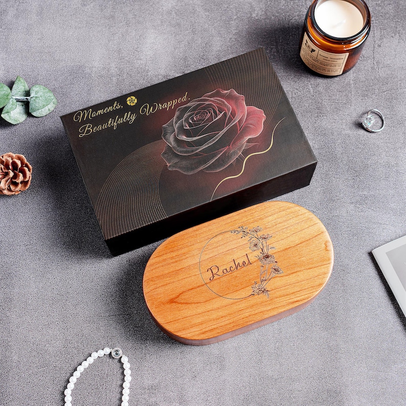May include: A rectangular black gift box featuring a red rose graphic and the text "Moments, Beautifully Wrapped." alongside an oval wooden box lid engraved with the name "Rachel" inside a circular floral wreath design on a gray surface.