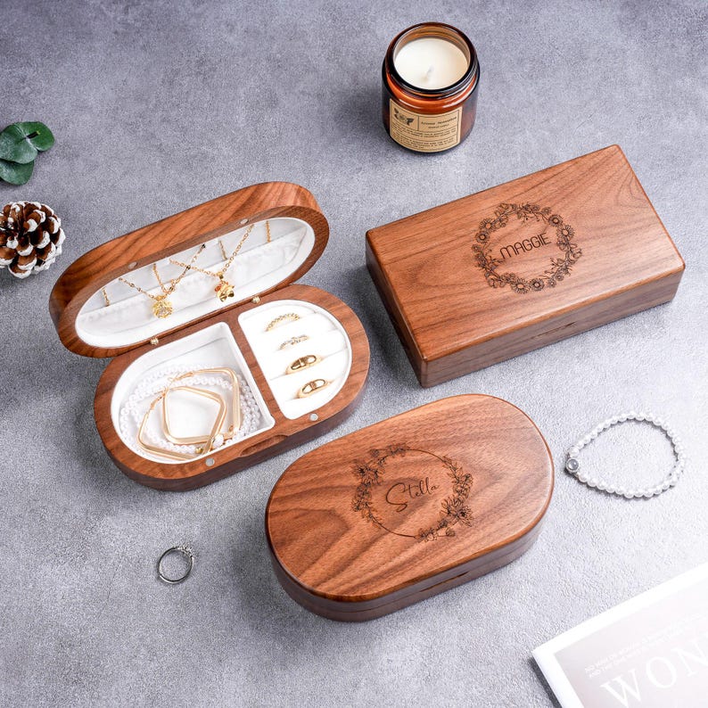 May include: Wooden jewelry boxes, one open to display necklaces, bracelets, and rings. The open box has a white interior. Two closed boxes feature floral wreath designs with names engraved. A lit candle and a bracelet are also present.