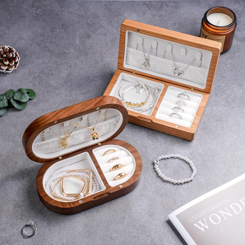 May include: Two wooden jewelry boxes, one oval and one rectangular, both open to display necklaces, bracelets, and rings. The boxes are lined with white fabric. A pearl bracelet and a candle are also visible.