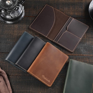 May include: A collection of leather passport holders in dark brown, navy blue, charcoal gray, tan, and forest green. One tan holder features the name Patrick embossed on the front. Each holder includes interior pockets for cards and travel documents.