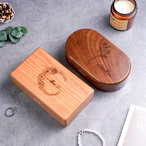 May include: Two wooden boxes with engraved designs. One is rectangular with a floral wreath and the name "Ethan" and a heart. The other is oval with a flower and the name "Amelia". Both are on a gray surface.