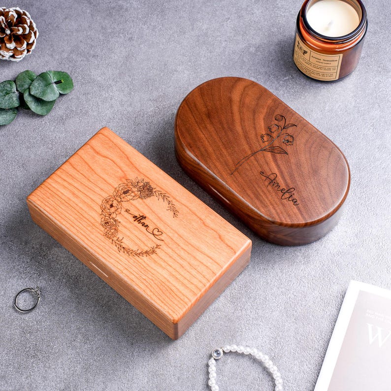 May include: Two wooden boxes with engraved designs. One is rectangular with a floral wreath and the name "Ethan" and a heart. The other is oval with a flower and the name "Amelia". Both are on a gray surface.