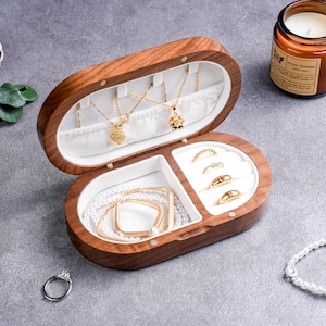 May include: An open, oval-shaped wooden jewelry box with a white interior. The box contains gold necklaces, rings, bracelets, and earrings. A lit candle sits in the background. The jewelry box is on a gray surface.