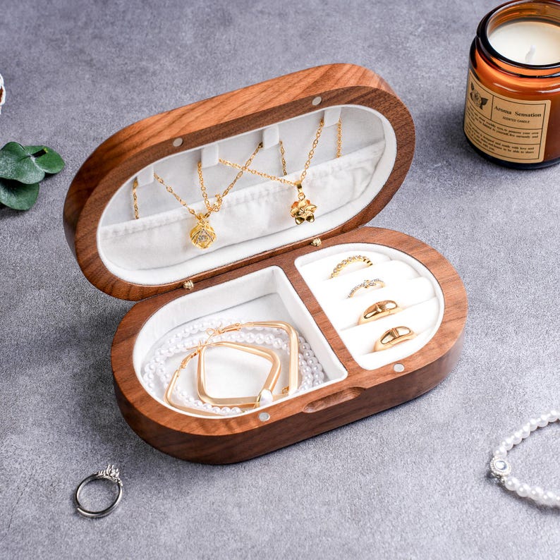 May include: An open, oval-shaped wooden jewelry box with a white interior. The box contains gold necklaces, rings, bracelets, and earrings. A lit candle sits in the background. The jewelry box is on a gray surface.