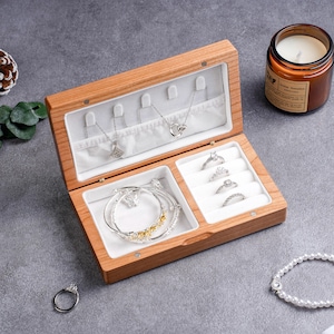 May include: Open wooden jewelry box with multiple compartments. The box contains necklaces, rings, and bracelets. The jewelry is silver and gold-toned. A lit candle in a glass jar is in the background.