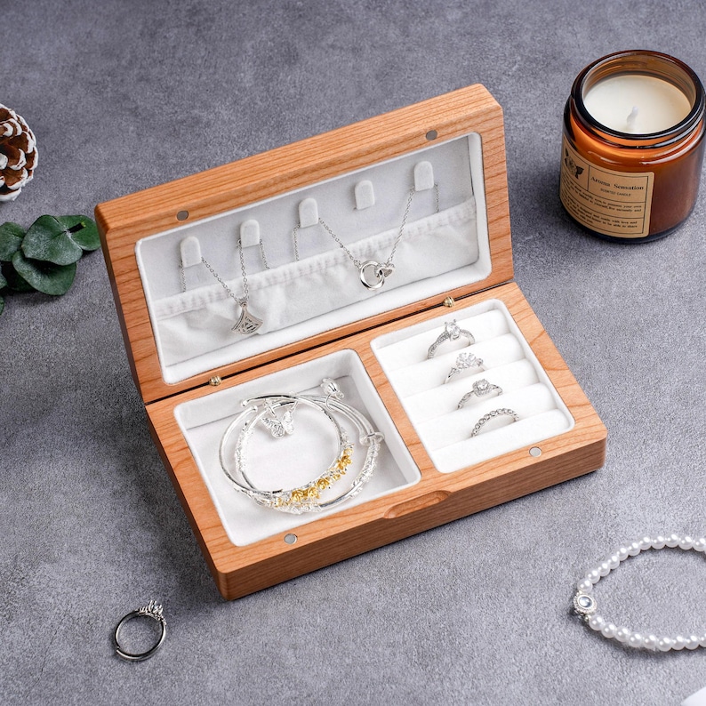May include: Open wooden jewelry box with multiple compartments. The box contains necklaces, rings, and bracelets. The jewelry is silver and gold-toned. A lit candle in a glass jar is in the background.