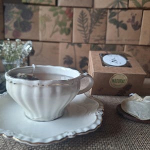 Könnte beinhalten: Eine weiße Teetasse und Untertasse mit goldenen Verzierungen, gefüllt mit Tee und einem Teebeutel. Ein kleiner Holzlöffel hält einen Teebeutel. Eine Teebox und dekorative Verpackungen sind im Hintergrund.